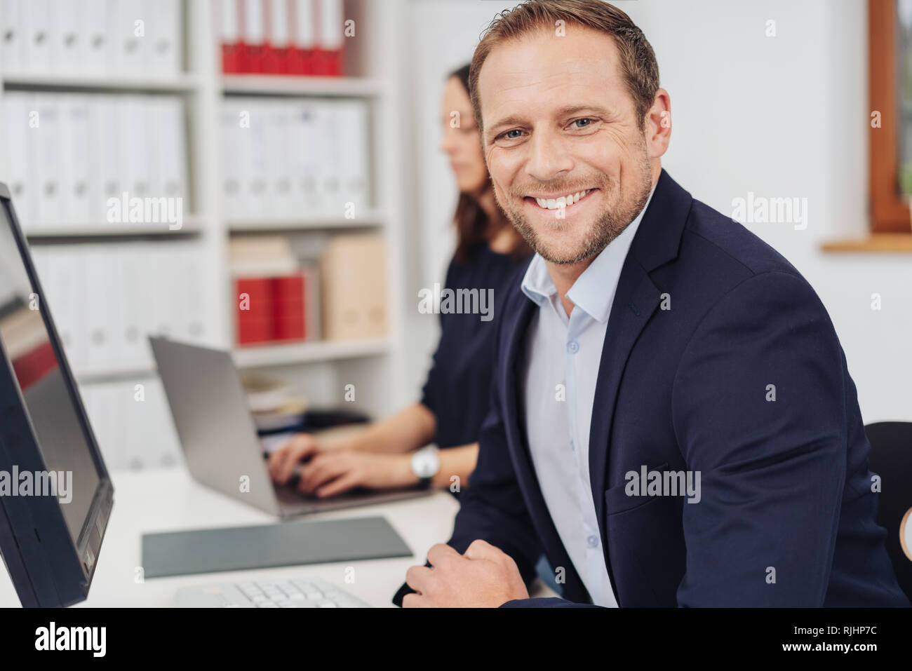 Beau et confident businessman in demi-tour portrait dans office interior, assis au bureau avec ordinateur, looking at camera et souriant. Son wom Banque D'Images