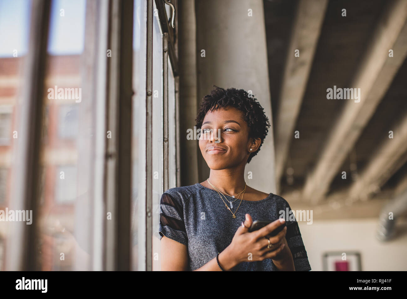 Young african american female looking out de fenêtre dans cet appartement loft holding smartphone Banque D'Images