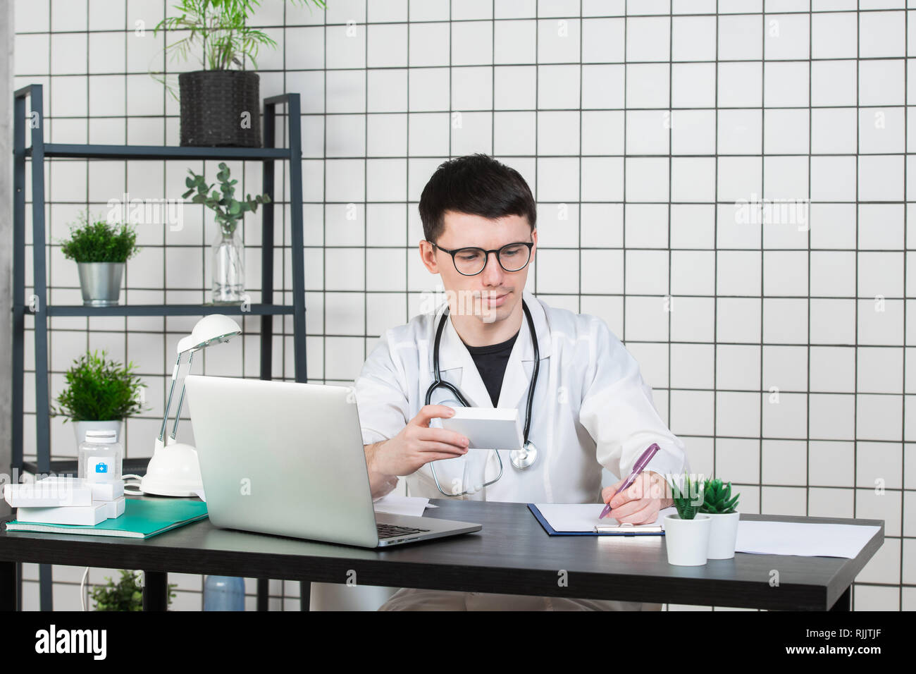 Médecin homme en blouse blanche avec stéthoscope sur son cou sitting at table pensant sur prescription, d'écrire quelque chose vers le bas, avec la boîte de médecine Banque D'Images