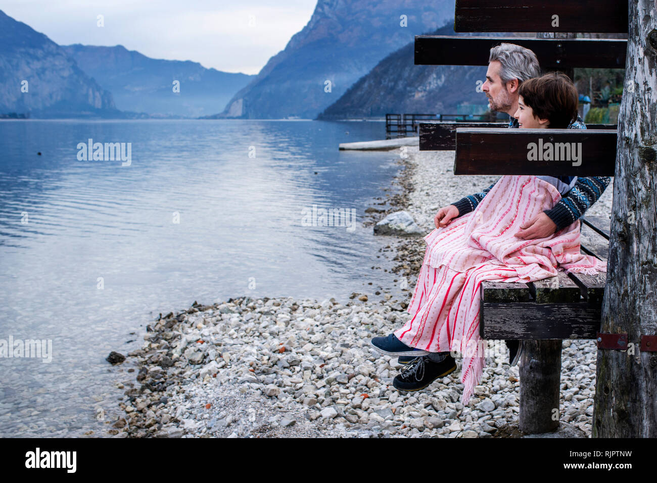 Garçon et père enveloppée de couverture sur la jetée au bord du lac, vue de côté, le lac de Côme, Onno, Lombardie, Italie Banque D'Images