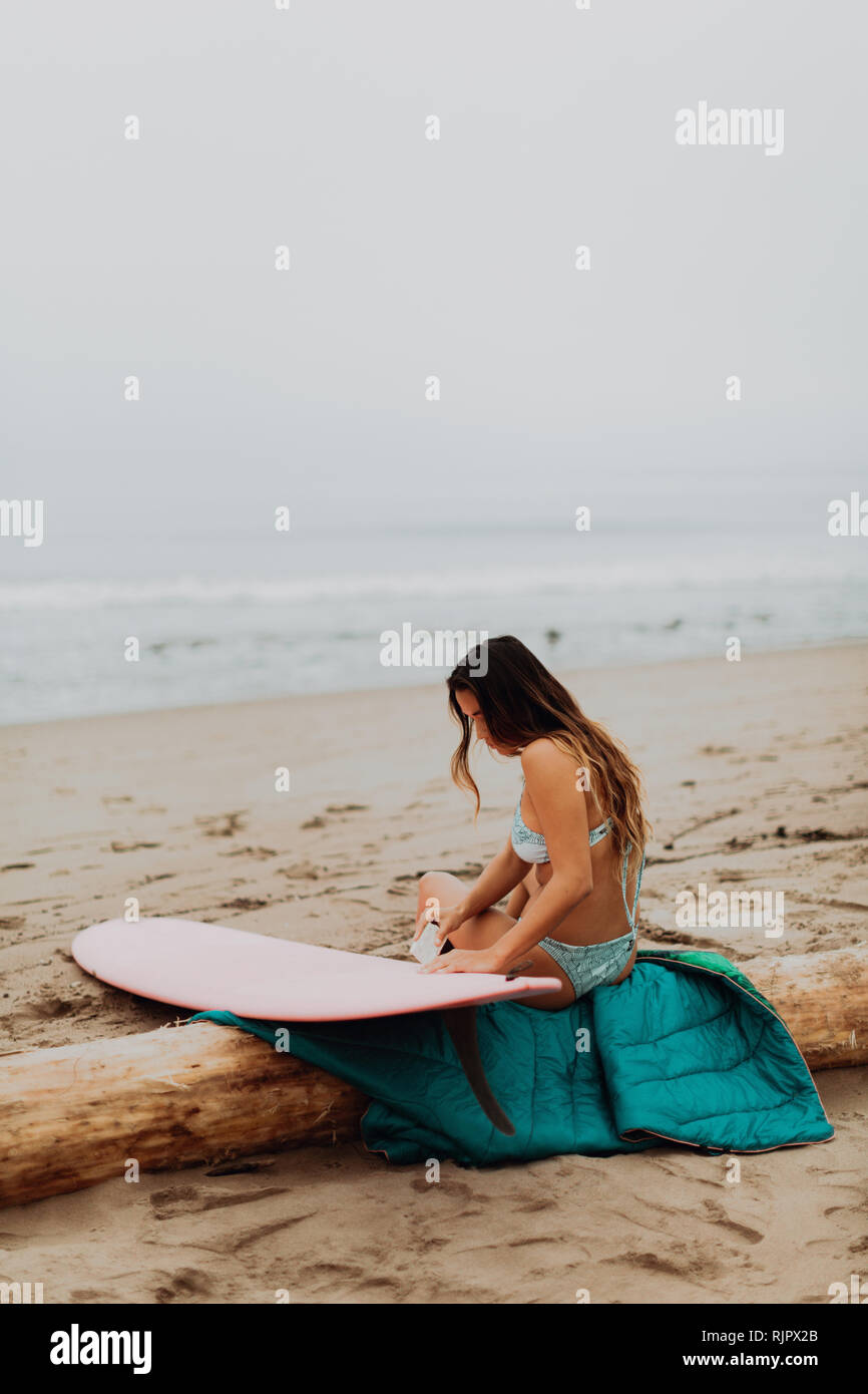Jeune femme surfer assis sur log waxing with surfboard on beach, Ventura, Californie, USA Banque D'Images
