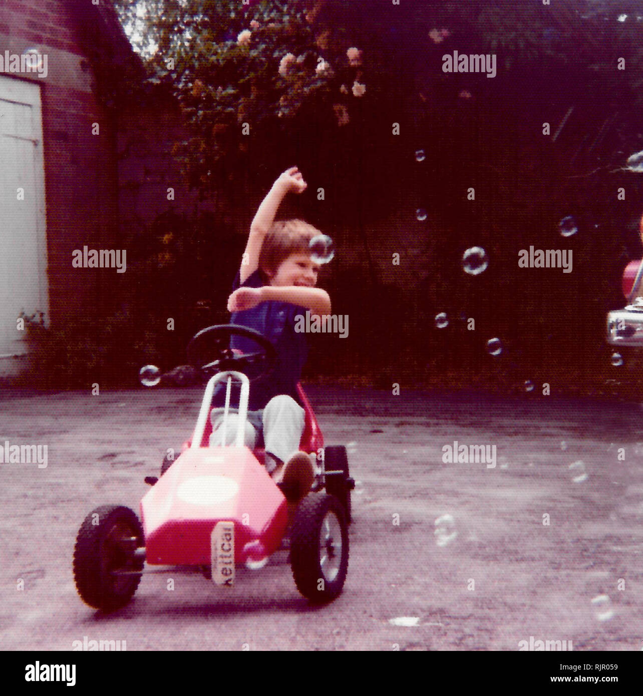 1970 - Un jeune garçon dans sa voiture à pédale zooms par bulles dans son jardin. Angleterre vers 1975. Banque D'Images