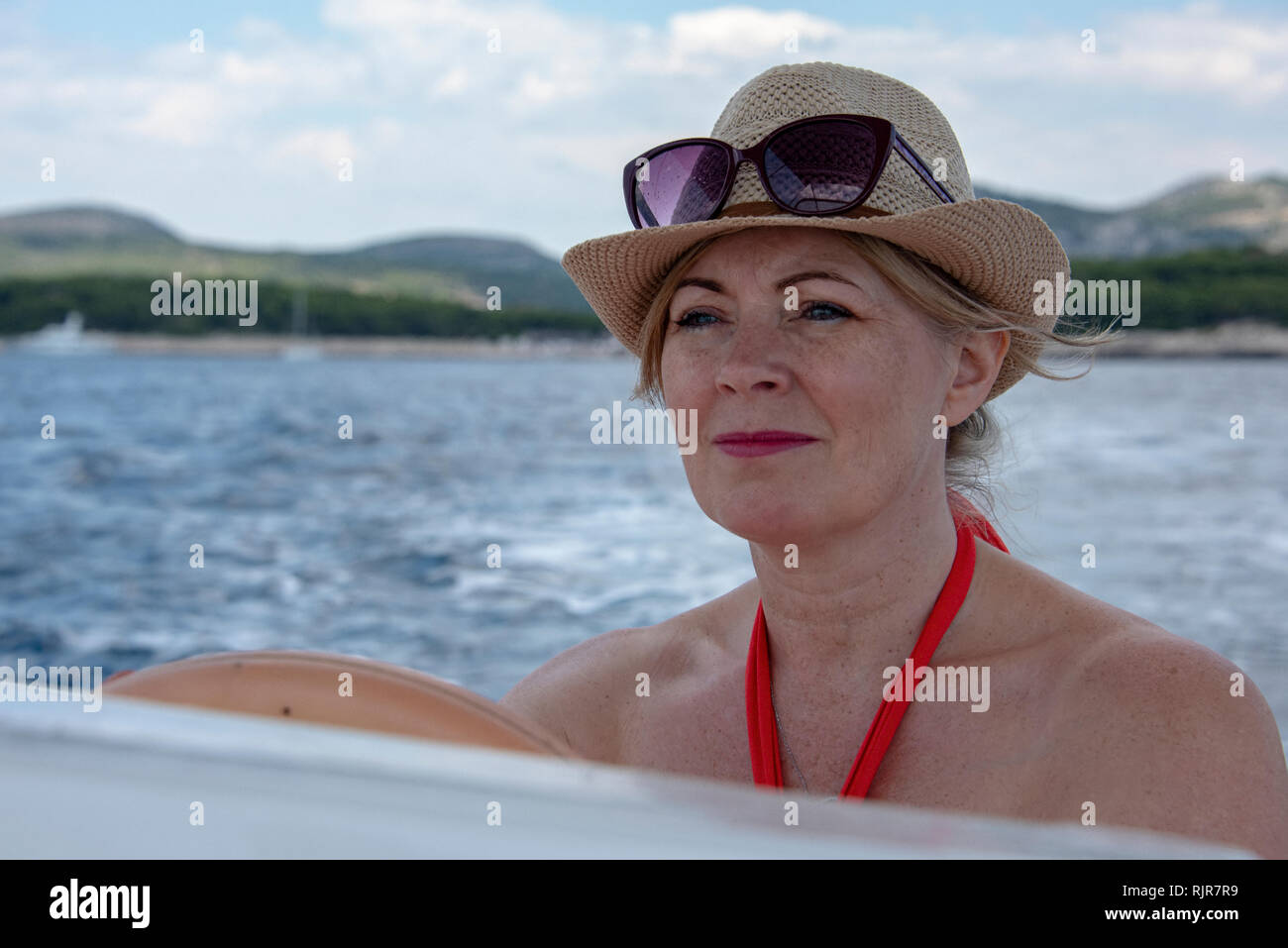 La Croatie, Hvar - Juin 2018 : entre 40 et 46 femmes dans la conduite d'un petit bateau. Elle porte une robe rouge et un chapeau chapeau et des lunettes de soleil sur sa tête. Littoral est visible Banque D'Images
