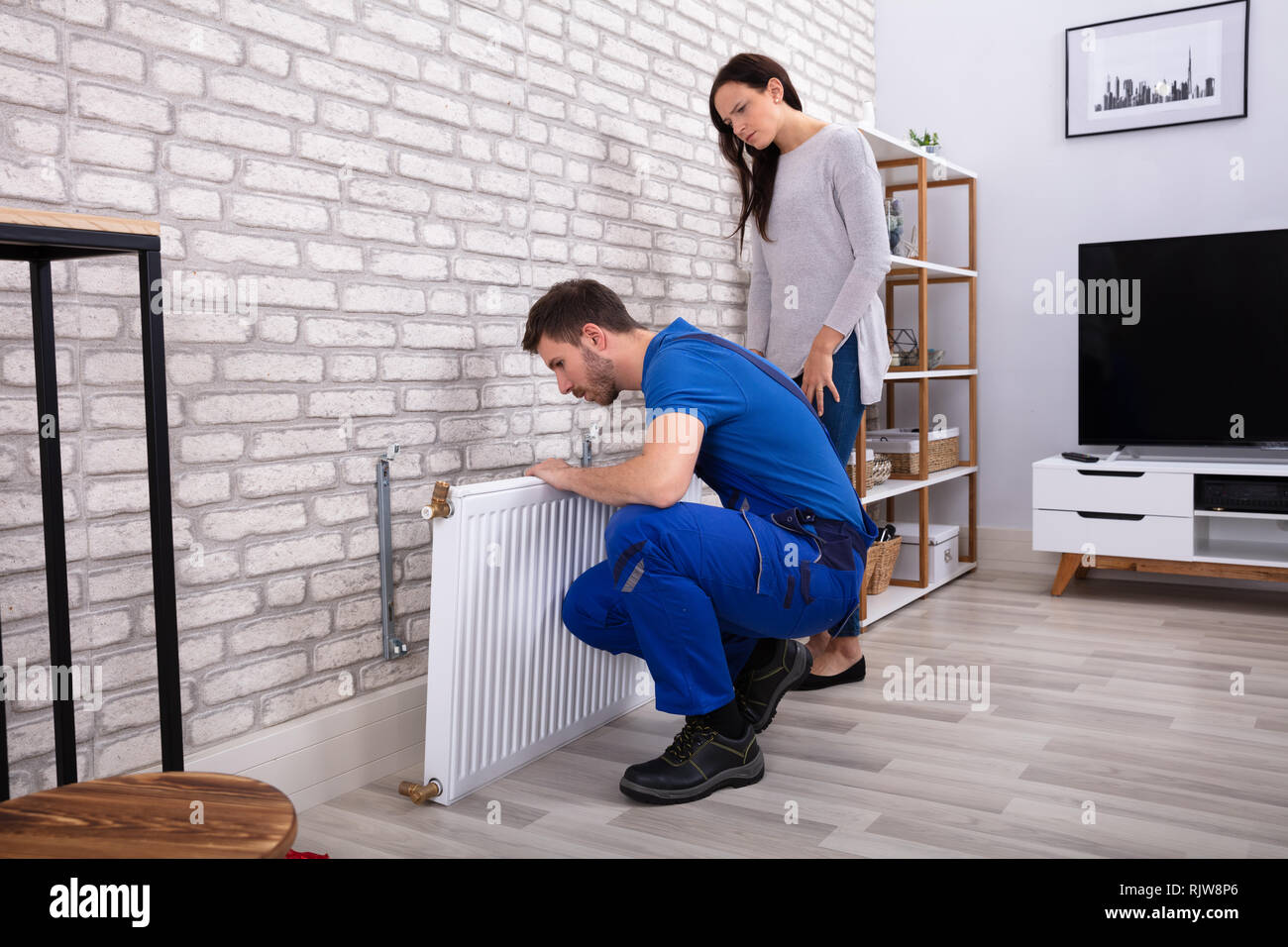 Jeune réparateur pose du radiateur sur le mur de briques avec femme debout à la maison Banque D'Images