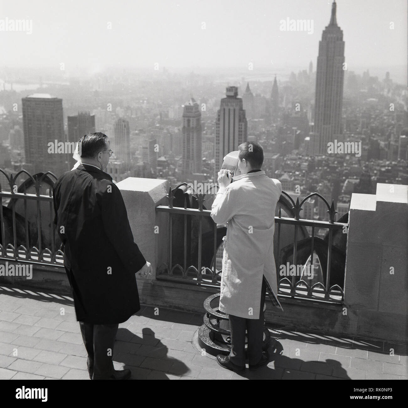 Années 1950, historique, à la recherche sur la ville de New York, New York, USA, deux hommes debout en haut d'un édifice en hauteur, l'un à l'aide d'un viewer ou télescope vue, avec l'Empire state immeuble au loin. Banque D'Images