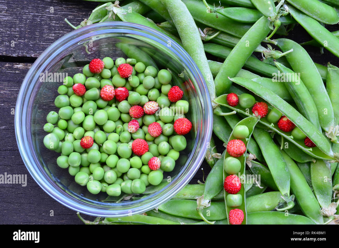 Pois écossés et fraises sauvages dans un bol en verre et quelques pods autour sur la vieille table en chêne, vue d'en haut Banque D'Images