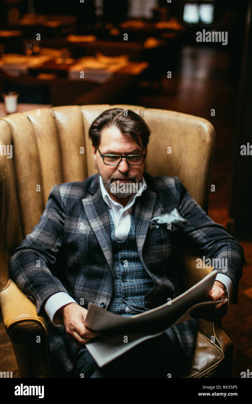 Portrait of mature succès caucasian Fashion designer élégant costume et portant des lunettes, la lecture de l'article à propos de sa nouvelle collection de vêtements en newspap Banque D'Images