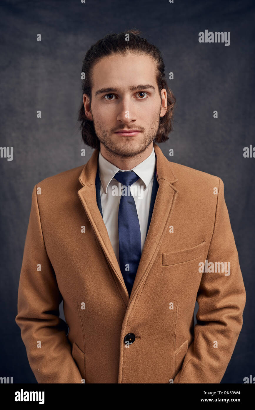 Portrait d'un jeune élégant bel homme aux longs cheveux brun clair non rasé en blazer et cravate bleu, looking at camera avec visage amical Banque D'Images