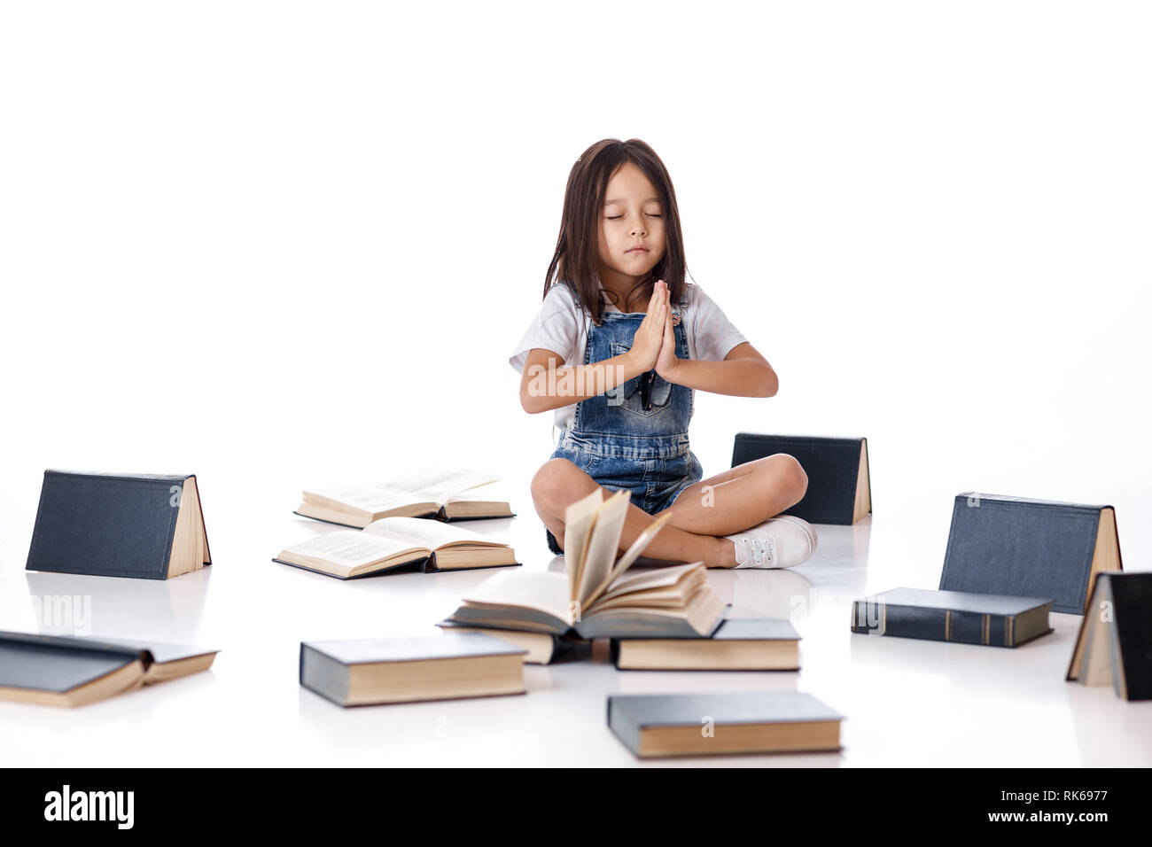 Cute little girl in denim assise dans une position du lotus entre les livres sur fond blanc. enfant et beaucoup de livres. Les enfants et l'éducation. Banque D'Images