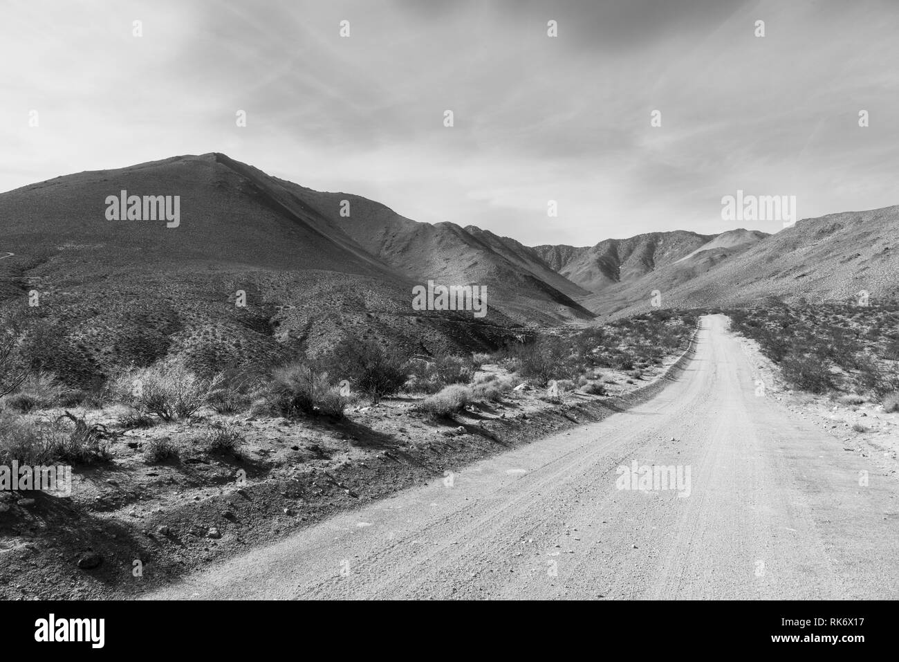 Noir et blanc, desert route de terre menant au désert aride montagne sous ciel nuageux. Banque de terre de chaque côté de la route et desert brosse. Banque D'Images