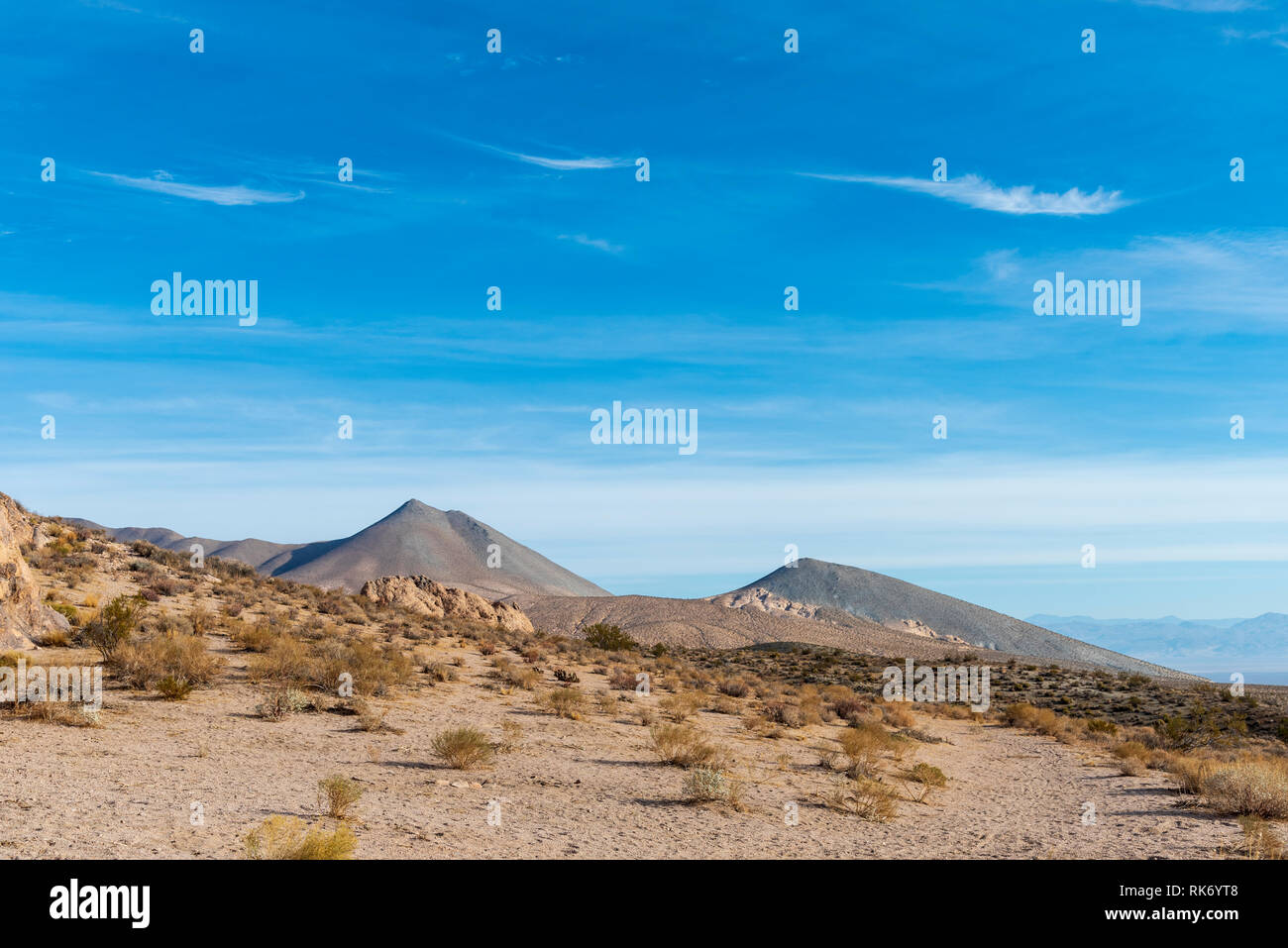 Colline du désert de sable avec des rochers et arbustes avec montagnes stériles au-delà sous ciel bleu avec des nuages. Banque D'Images