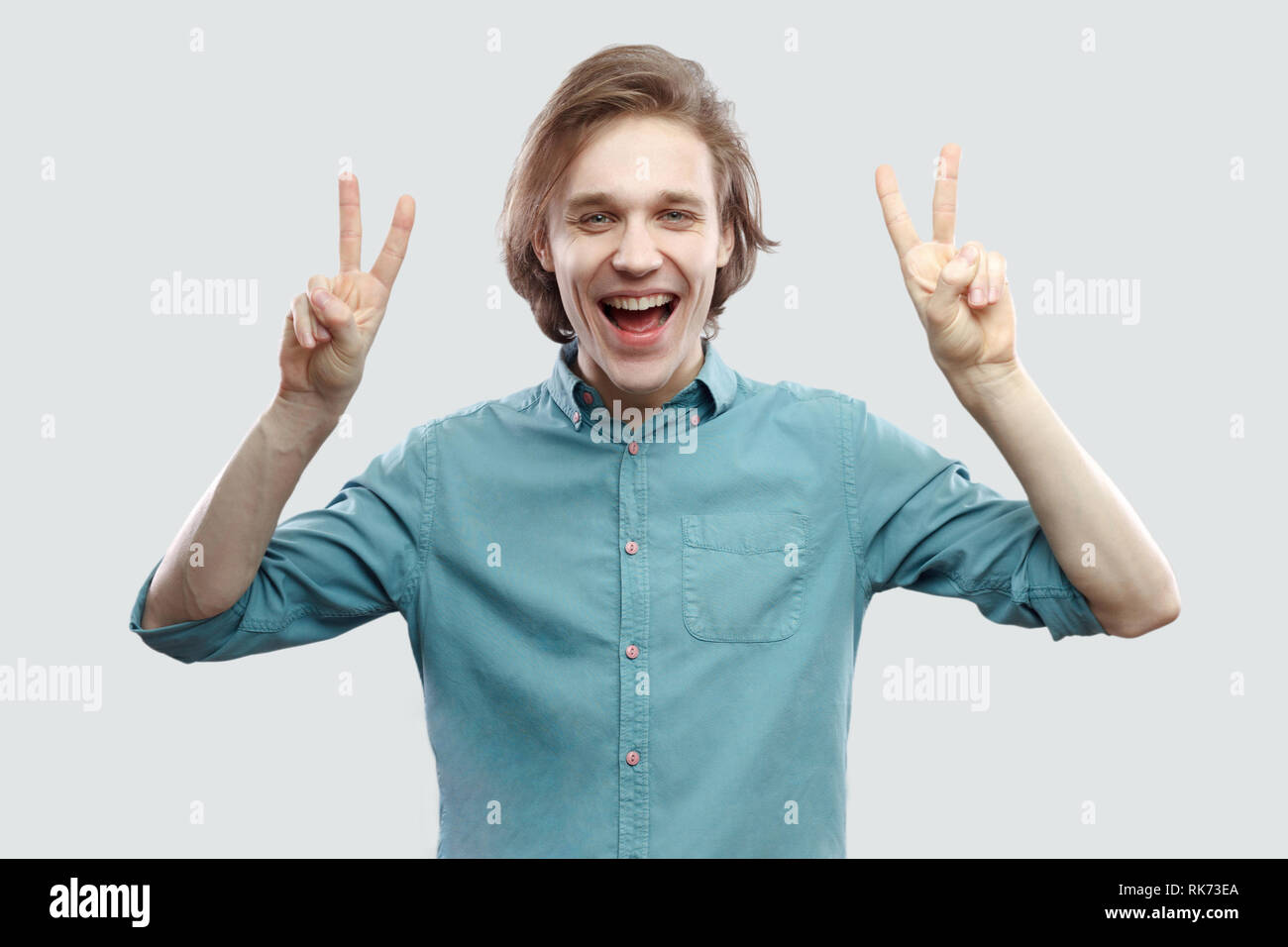 Portrait of happy beau jeune homme blond aux cheveux longs en chemise bleu et permanent à la caméra au visage, avec une victoire ou la paix indo geste. Banque D'Images