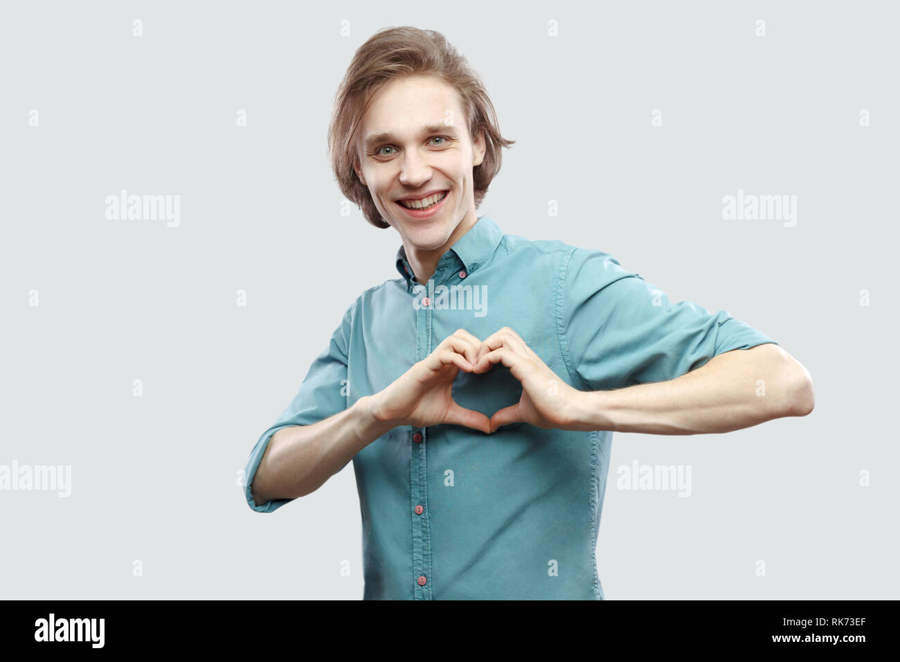 Portrait of happy beau jeune homme blond aux cheveux longs en bleu chemise décontractée, sourire permanent à pleines dents, coeur d'amour avec des gestes rugueux sourire intérieur. Banque D'Images