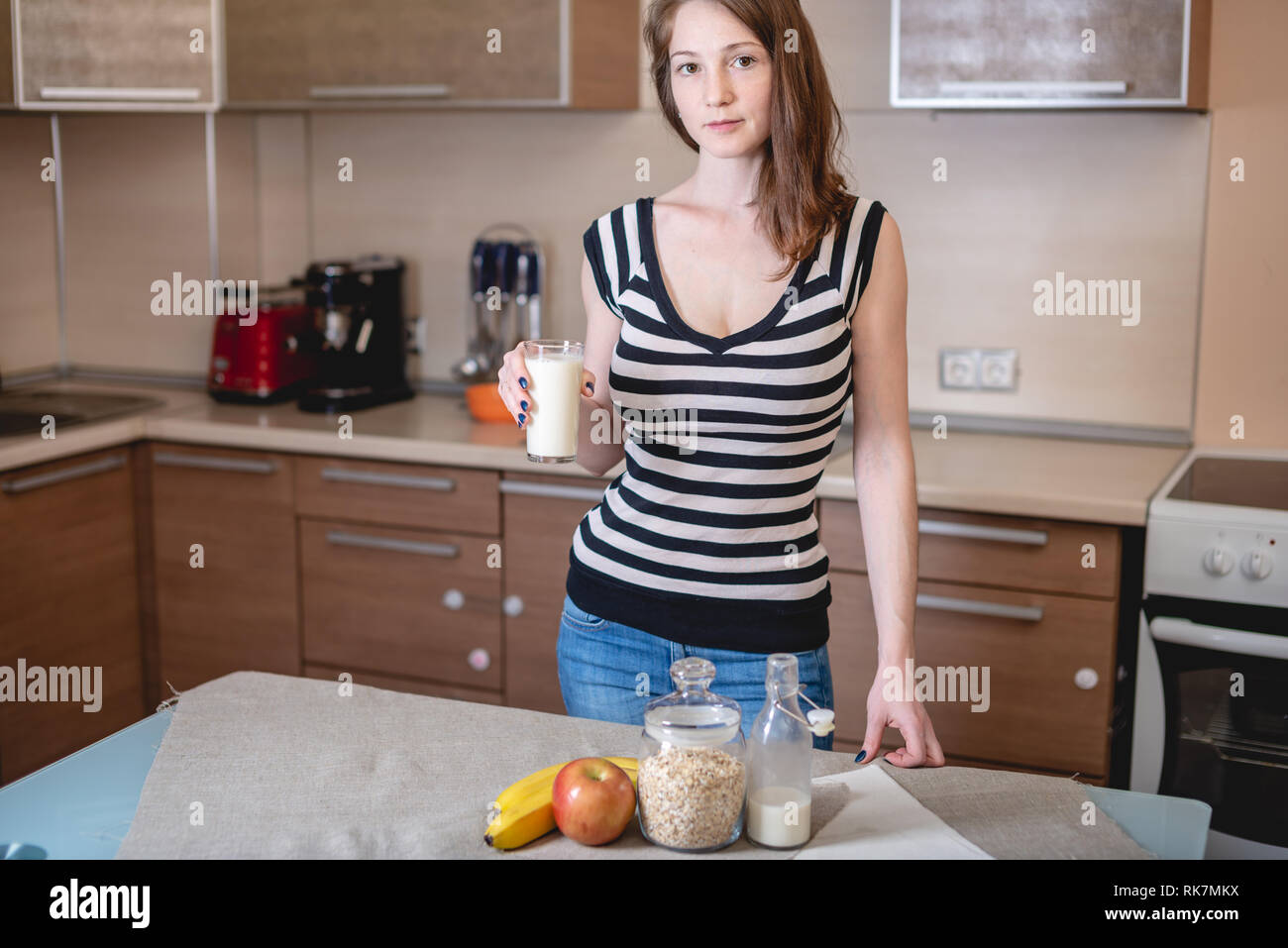 Jeune femme tenant un verre de lait d'avoine bio dans la cuisine. Fruits et bouteille sur la table. Produit végétarien sain de régime Banque D'Images