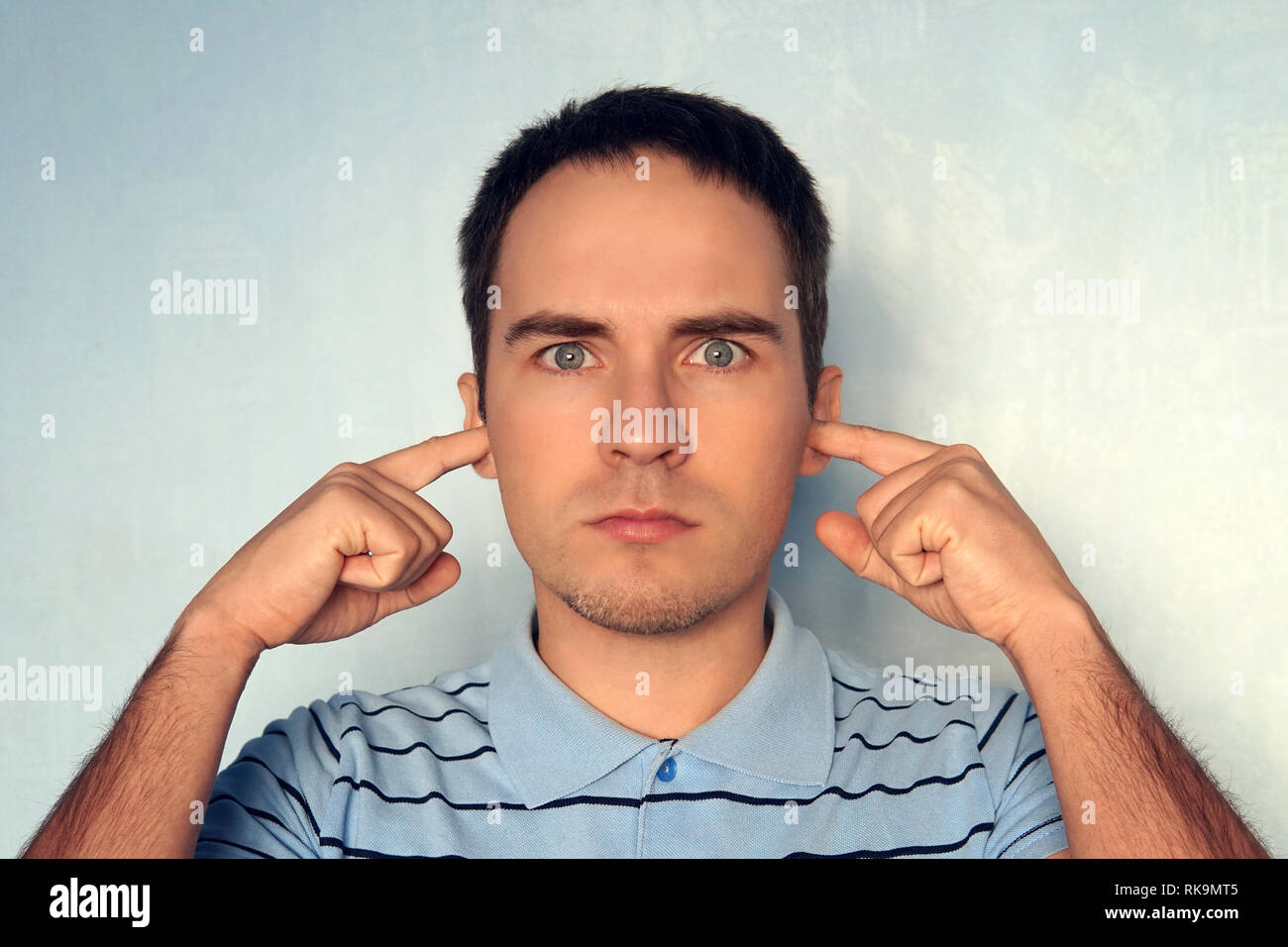 Beau jeune homme mécontent avec oreilles bouchons modèle expression comme entend le bruit fort ennuyeux ou roaring non loin de lui, plus de mur de béton bleu. Pas Banque D'Images