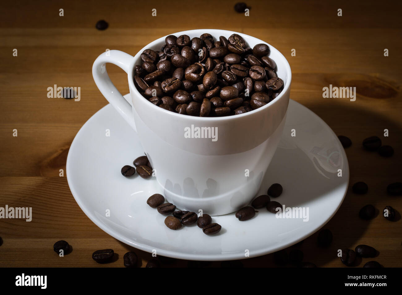 Tasse blanche remplie de grains de café et café en grains éparpillés sur une surface en bois et sur la soucoupe blanche. Banque D'Images