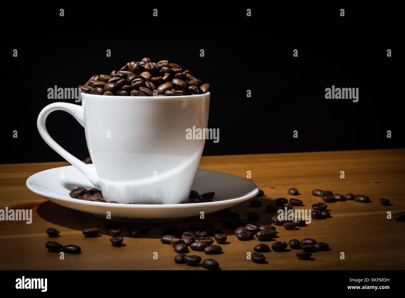Tasse blanche remplie de grains de café et café en grains éparpillés sur une surface en bois et sur la soucoupe blanche. Banque D'Images