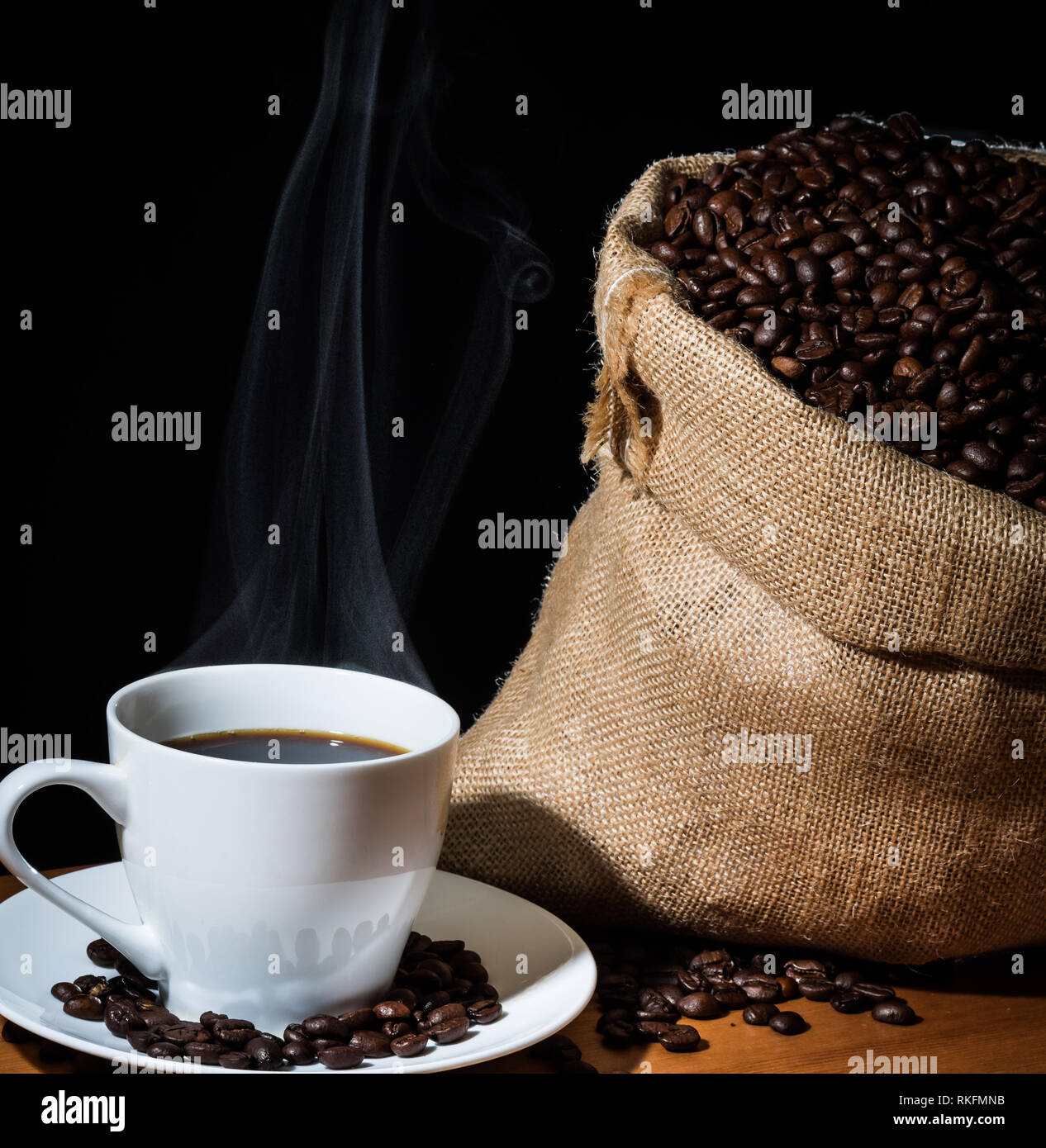 Café infusé dans une tasse blanche avec de la vapeur ou de la vapeur et les grains de café dans une toile de jute sac en toile de jute et les haricots éparpillés sur une surface en bois. Banque D'Images