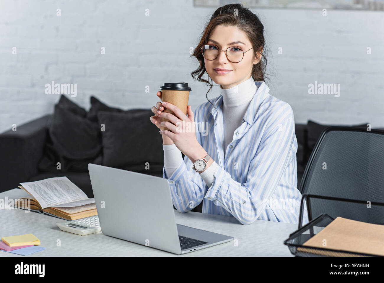 Attractive businesswoman holding paper cup, using laptop and looking at camera Banque D'Images