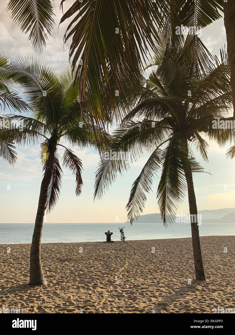 Homme seul s'assied sur la plage de sable près de sa moto et s'intéresse à la surface de la mer sur le fond du ciel bleu avec plusieurs nuages. Les palmiers d'un vert Banque D'Images