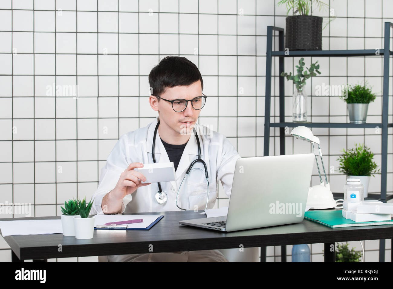 Médecin homme en blouse blanche avec stéthoscope sur son cou sitting at table pensant sur prescription, d'écrire quelque chose vers le bas, avec la boîte de médecine Banque D'Images