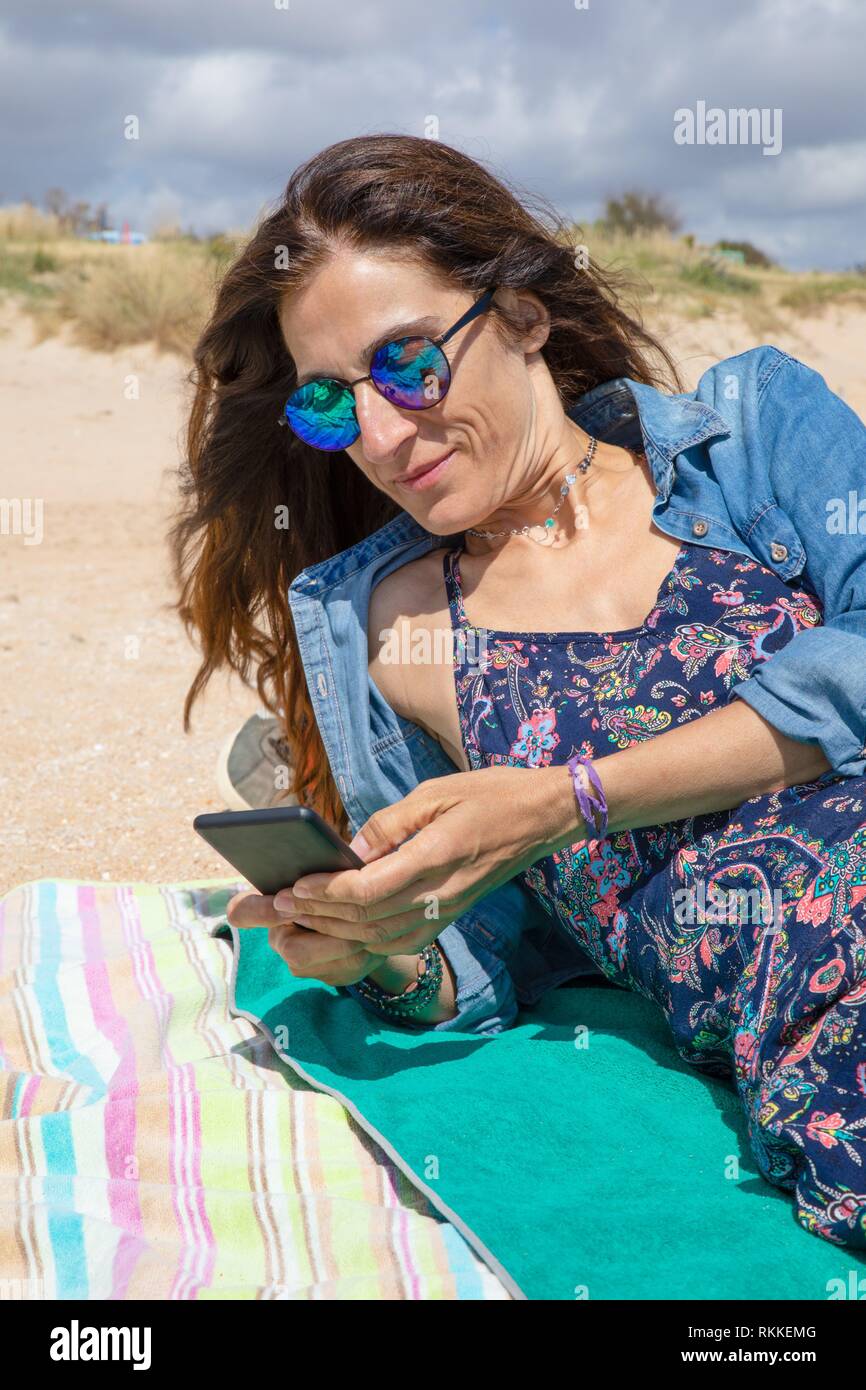 Femme brune avec des lunettes de soleil et des vêtements bleus de sourire  et de toucher pour téléphone intelligent ou mobile, SMS, allongé sur une  serviette sur la plage Photo Stock -