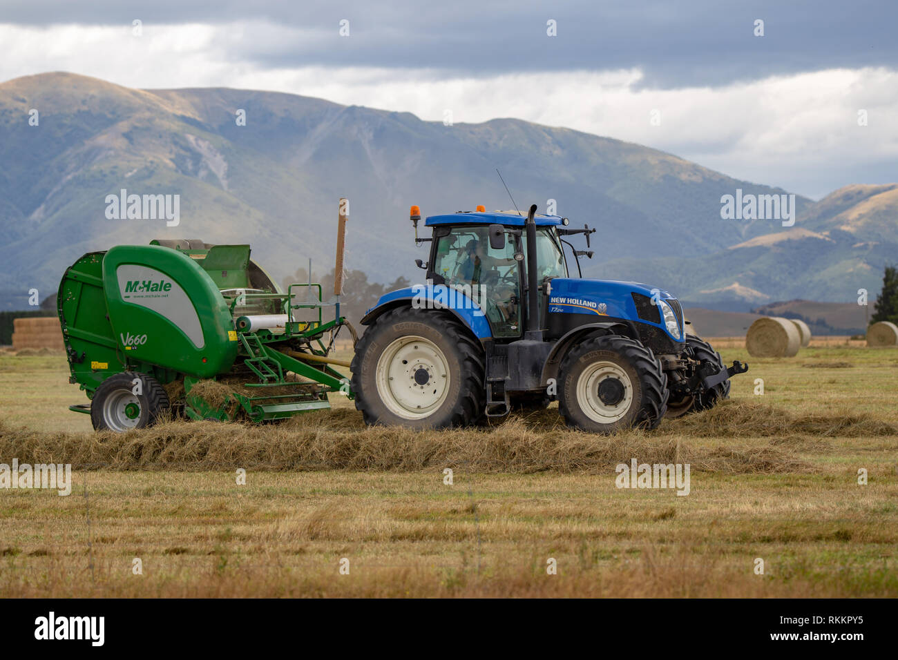 Annat, Canterbury, Nouvelle-Zélande - 1 Février 2019 : Un tracteur bleu avec une presse à balles de foin travaille son chemin le long des rangées de balles rondes dans un domaine rural Banque D'Images