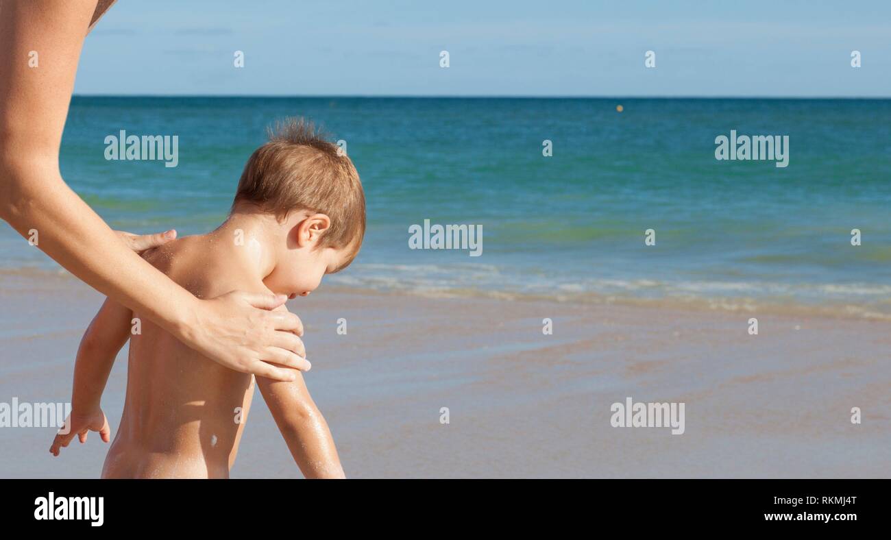 L'application de crème solaire à la mère de son petit fils sur le dos. Fond de plage idyllique. Banque D'Images