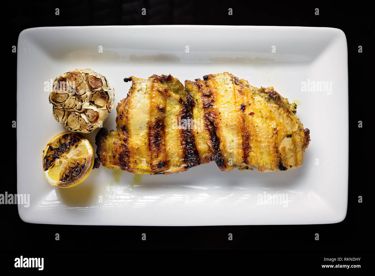 Photo du haut vers le bas d'un blanc de poulet grillé servi dessus une assiette blanche avec de l'ail cuit et du citron Banque D'Images