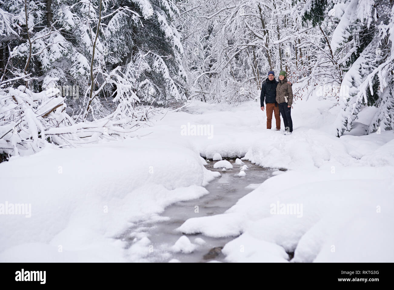 Smiling young couple in hiking gear standing by a partiellement congelée rivière qui traverse une forêt couverte de neige tandis que dehors pour une randonnée d'hiver Banque D'Images