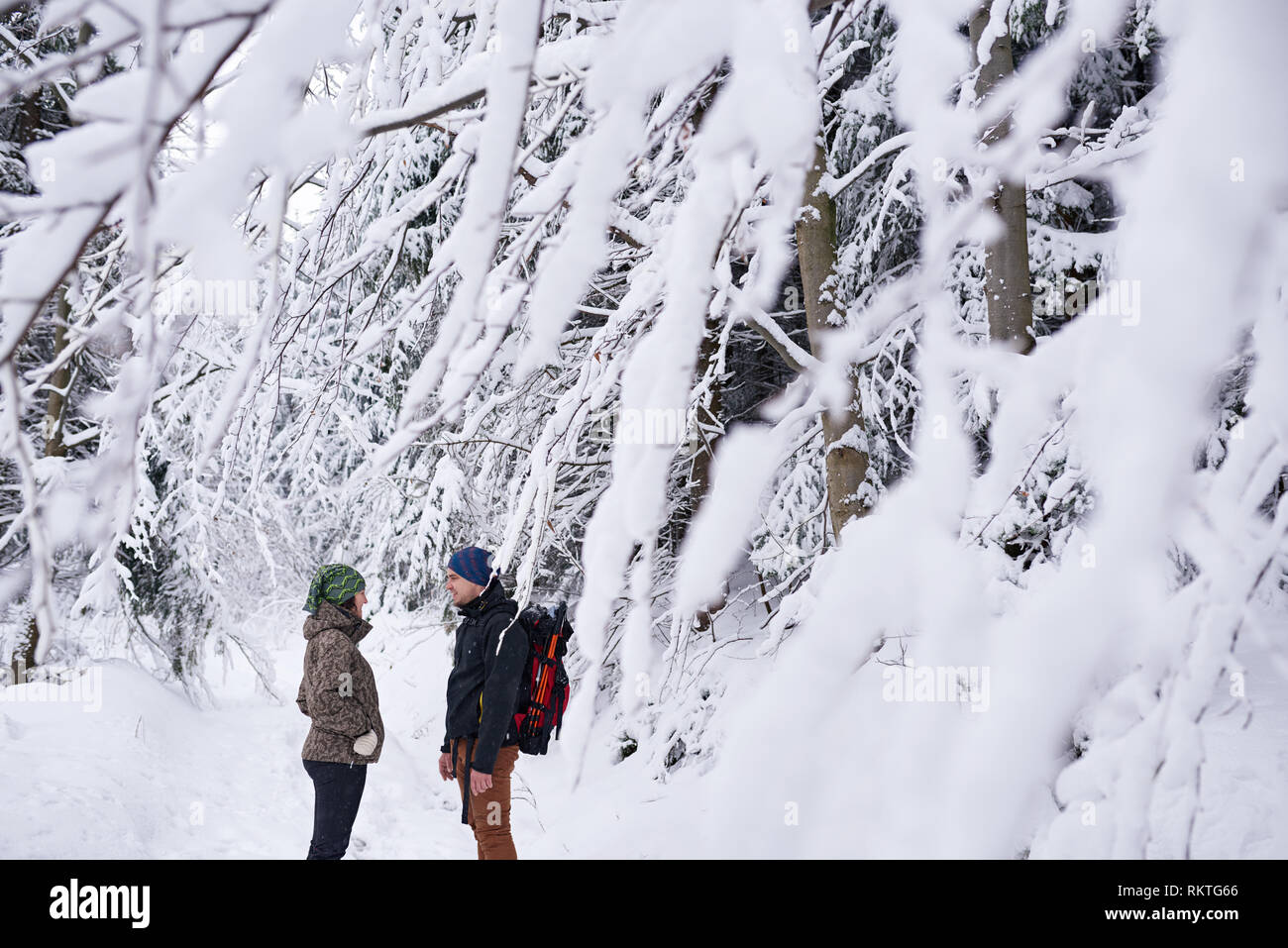 Smiling young couple en parlant ensemble d'équipement de randonnée en étant debout sur un chemin dans une forêt couverte de neige pendant une randonnée d'hiver Banque D'Images