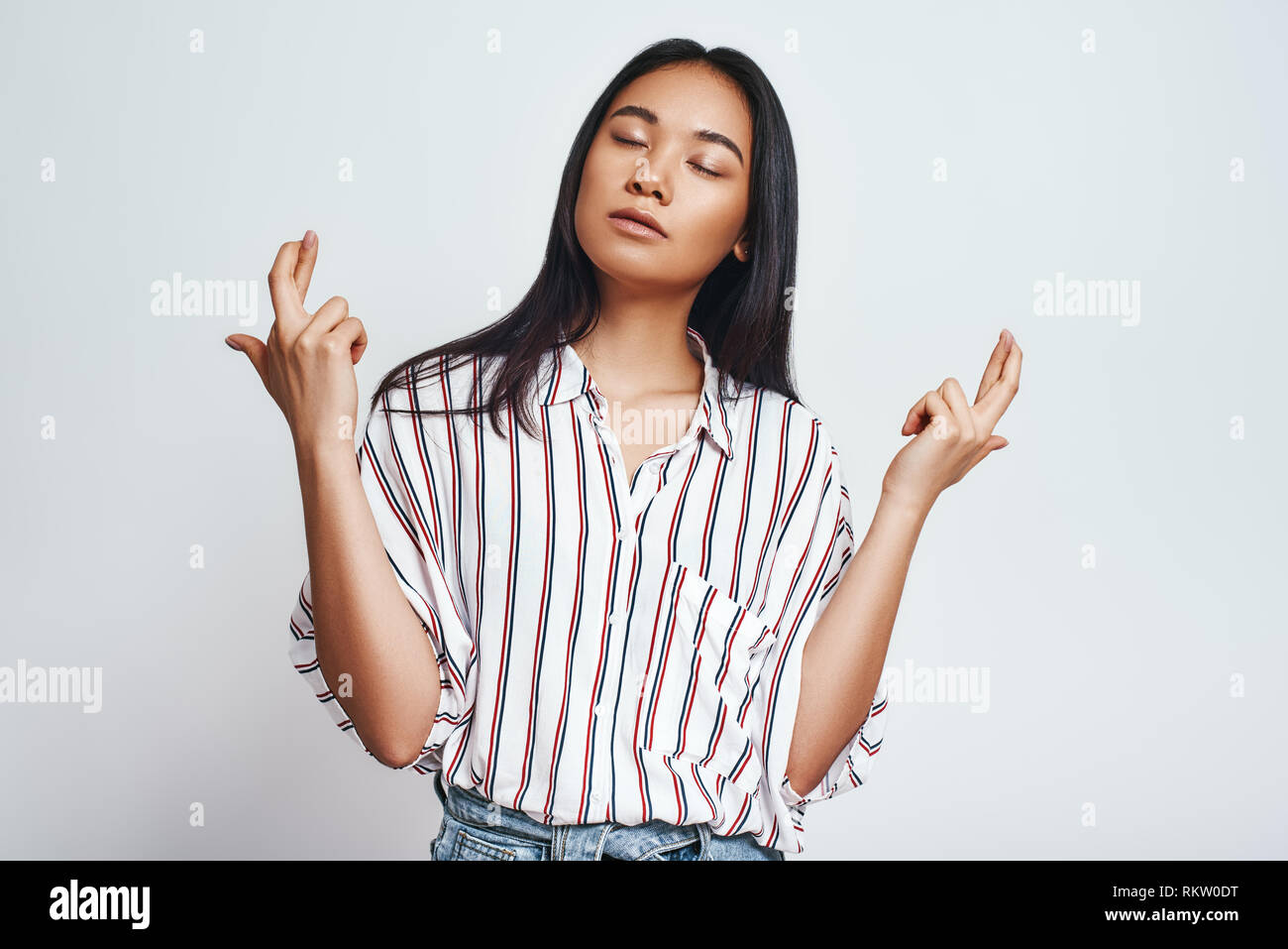 En attente d'un moment unique. Portrait de jeune femme asiatique de rêve dans des vêtements décontractés crossing fingers et penser à son souhait en étant debout sur un fond gris. Wishness concept. Banque D'Images