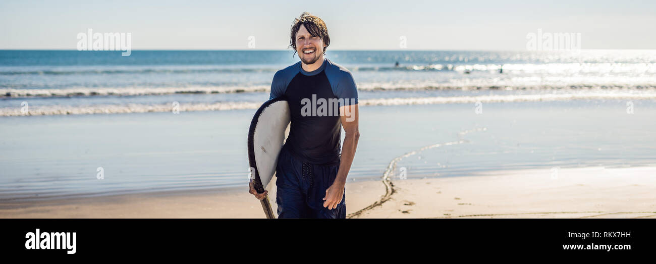 Beau jeune sportif posant avec sa planche de surf sous le bras dans sa combinaison sur une plage tropicale de sable long format bannière Banque D'Images