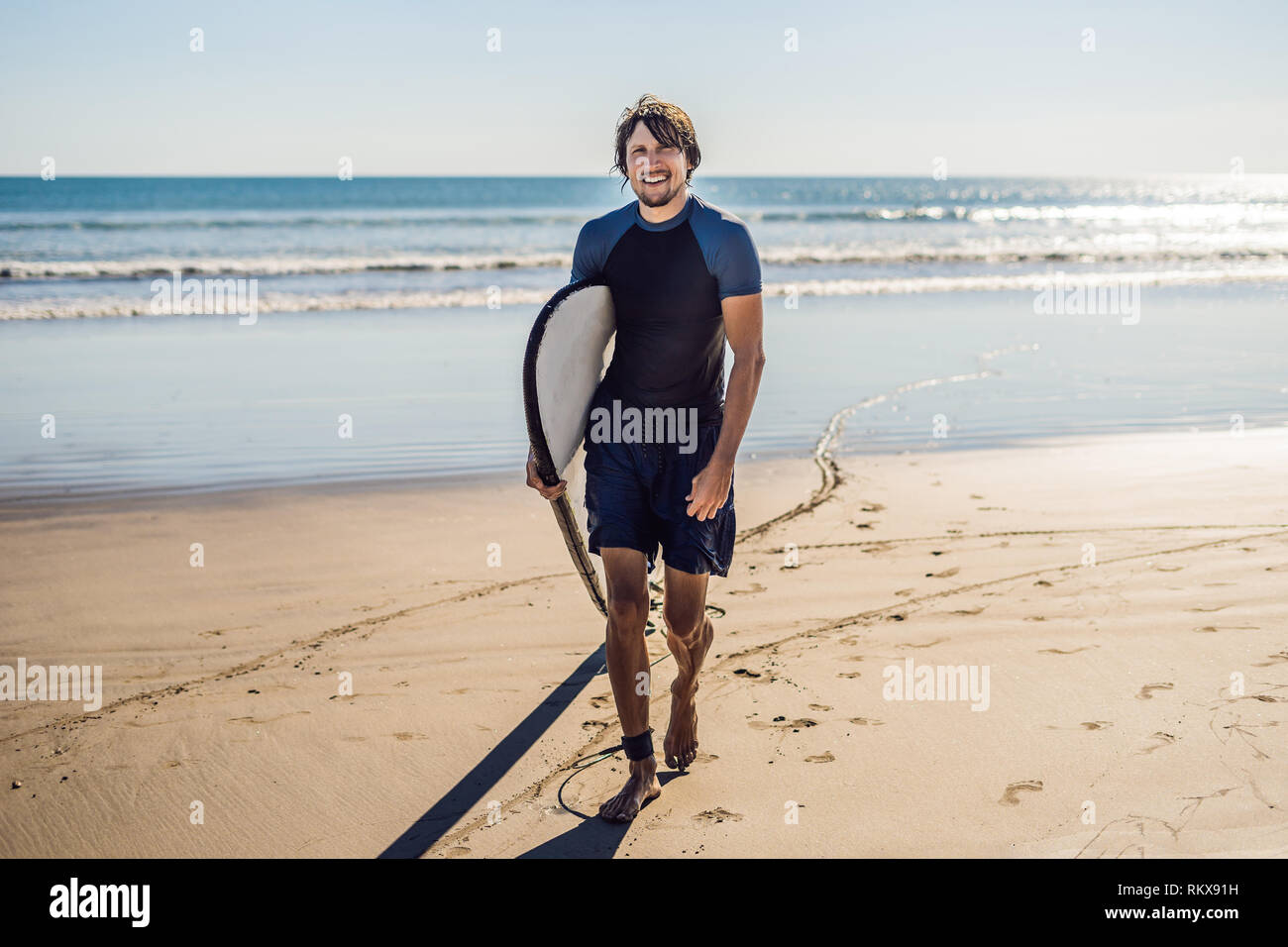 Beau jeune sportif posant avec sa planche de surf sous le bras dans sa combinaison sur une plage tropicale de sable Banque D'Images