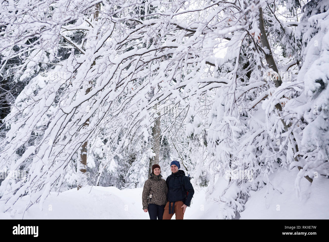 Portrait of a smiling young couple in hiking gear standing arm in arm ensemble sur un sentier de la forêt enneigée en hiver Banque D'Images