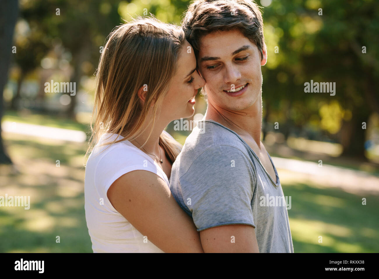 Romantic couple standing in park passer du temps ensemble. Woman hugging son petit ami par derrière et de toucher leurs têtes debout à l'extérieur. Banque D'Images