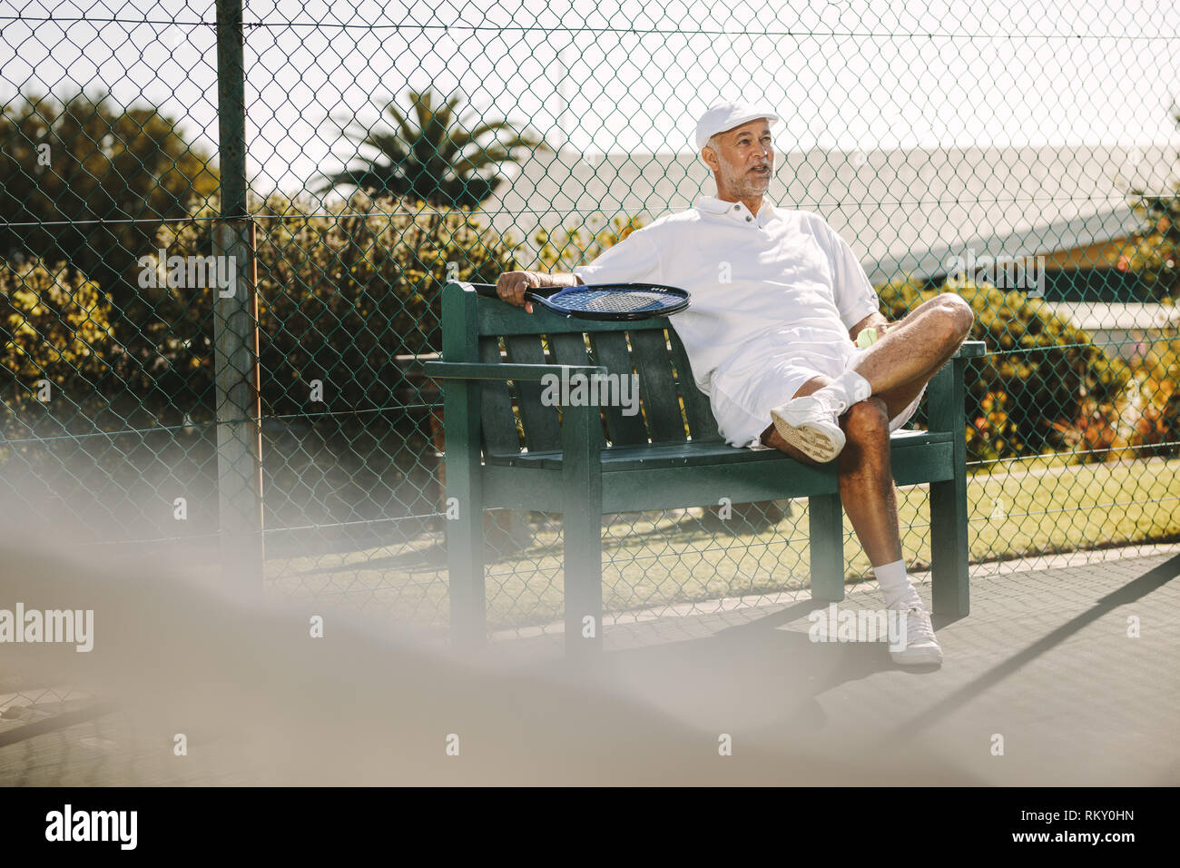 Personne âgée assise sur un banc à côté d'un court de tennis et vous détendre. Dans l'homme de prendre l'usure tennis reste pendant une partie de tennis. Banque D'Images