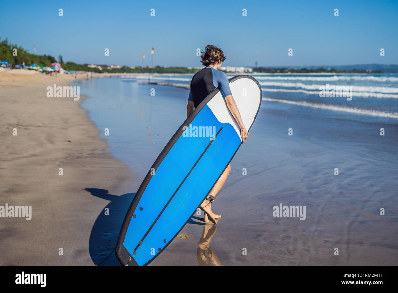 Beau jeune sportif posant avec sa planche de surf sous le bras dans sa combinaison sur une plage tropicale de sable Banque D'Images