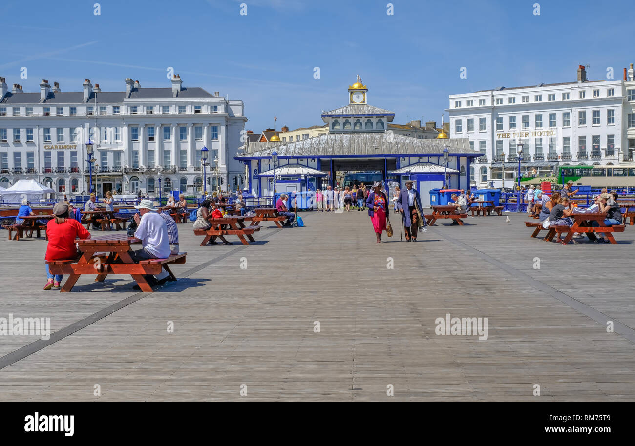 Eastbourne, Sussex, England, UK - 1 août 2018 : les uns flânant et assis sur des bancs de pique-nique près de l'entrée de la jetée, à Eastbourne. Le Cla Banque D'Images