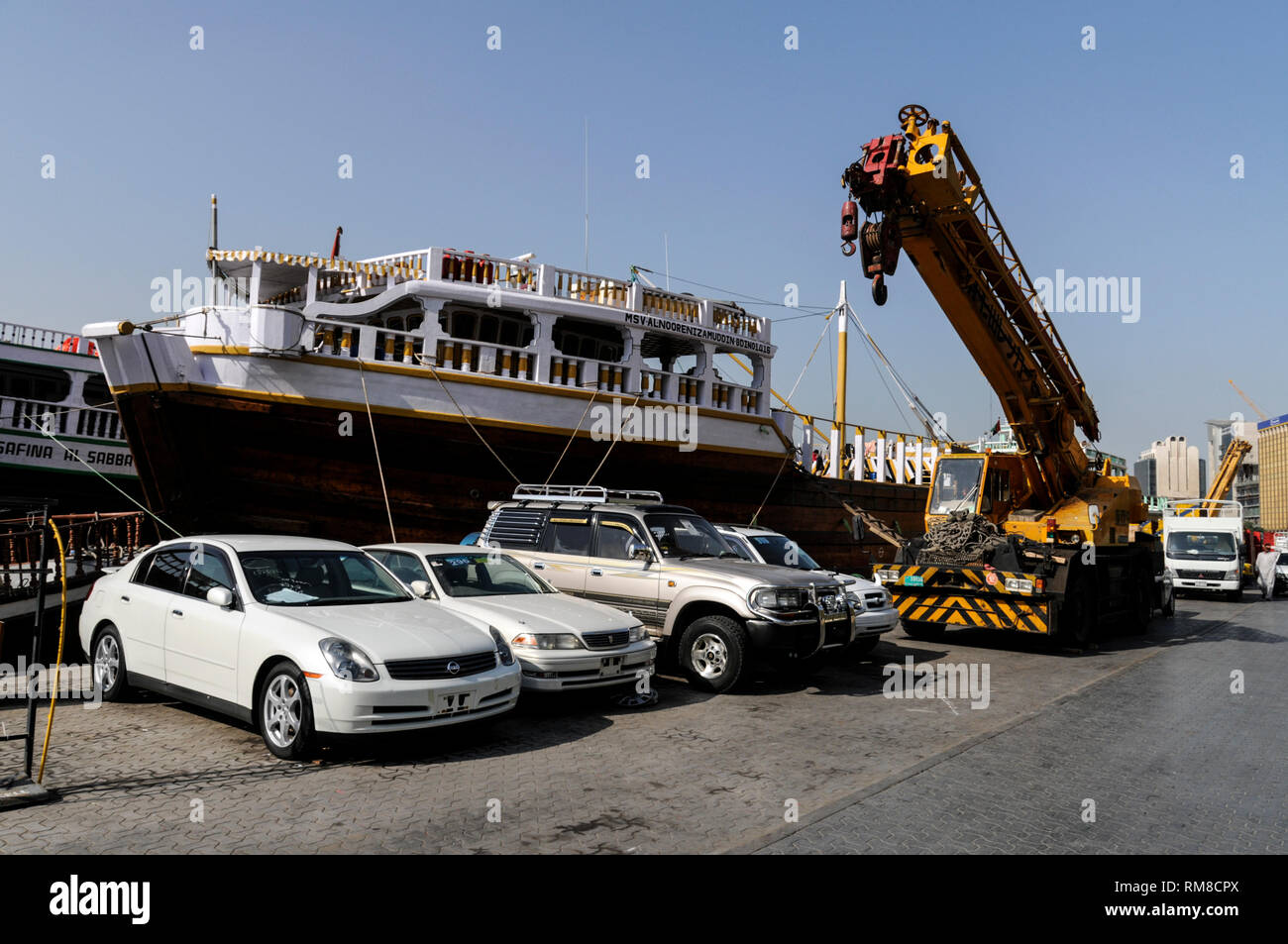 Une flotte de véhicules en attente d'être hébergé par comme grue pour être chargés à bord d'un boutre en bois de haute mer ( Bateau Arabe) d'être chargé de fret sur le Dubai C Banque D'Images