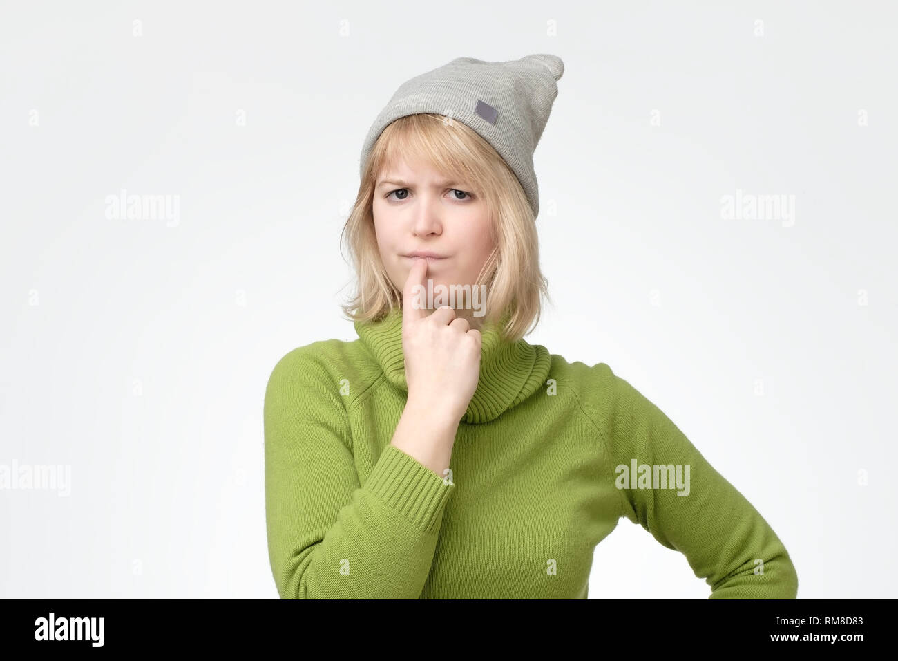 Femme blonde wearing green pullover avec expression perplexe, maintient le doigt sur les lèvres Banque D'Images