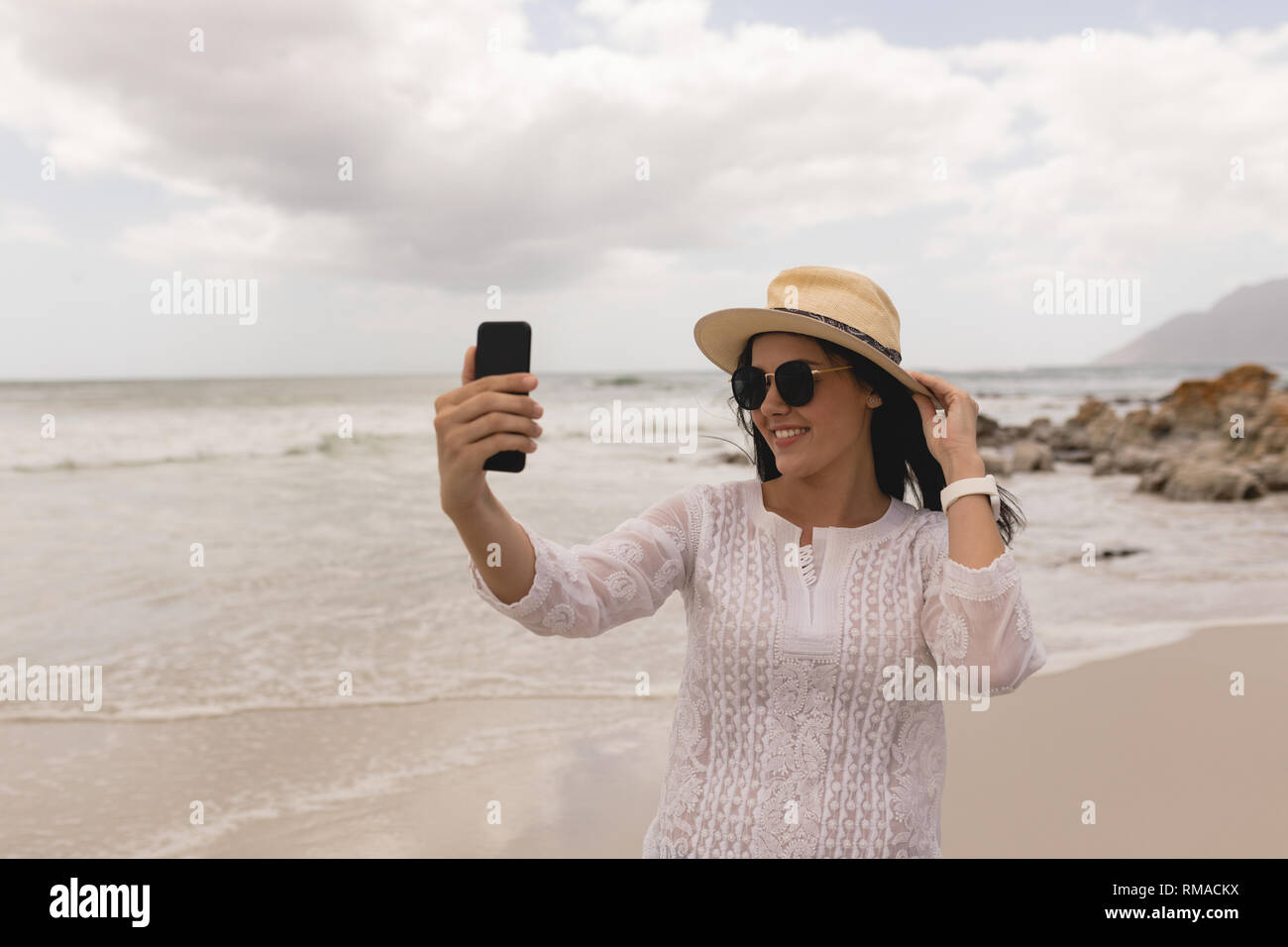 Jeune femme avec chapeau et des lunettes en tenant avec selfies mobile phone on beach Banque D'Images