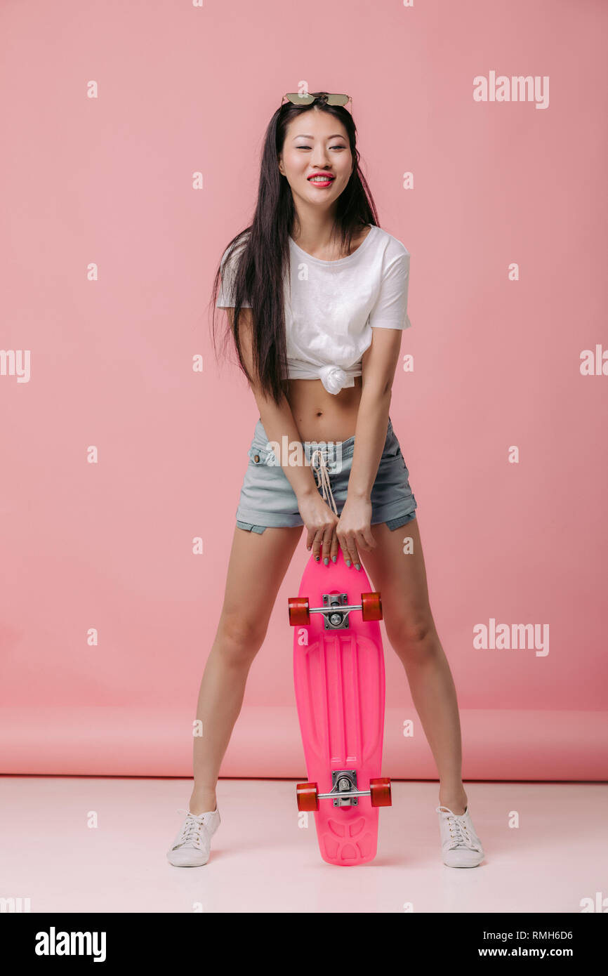 Beautiful smiling asian woman in sunglasses holding skateboard sur fond rose Banque D'Images