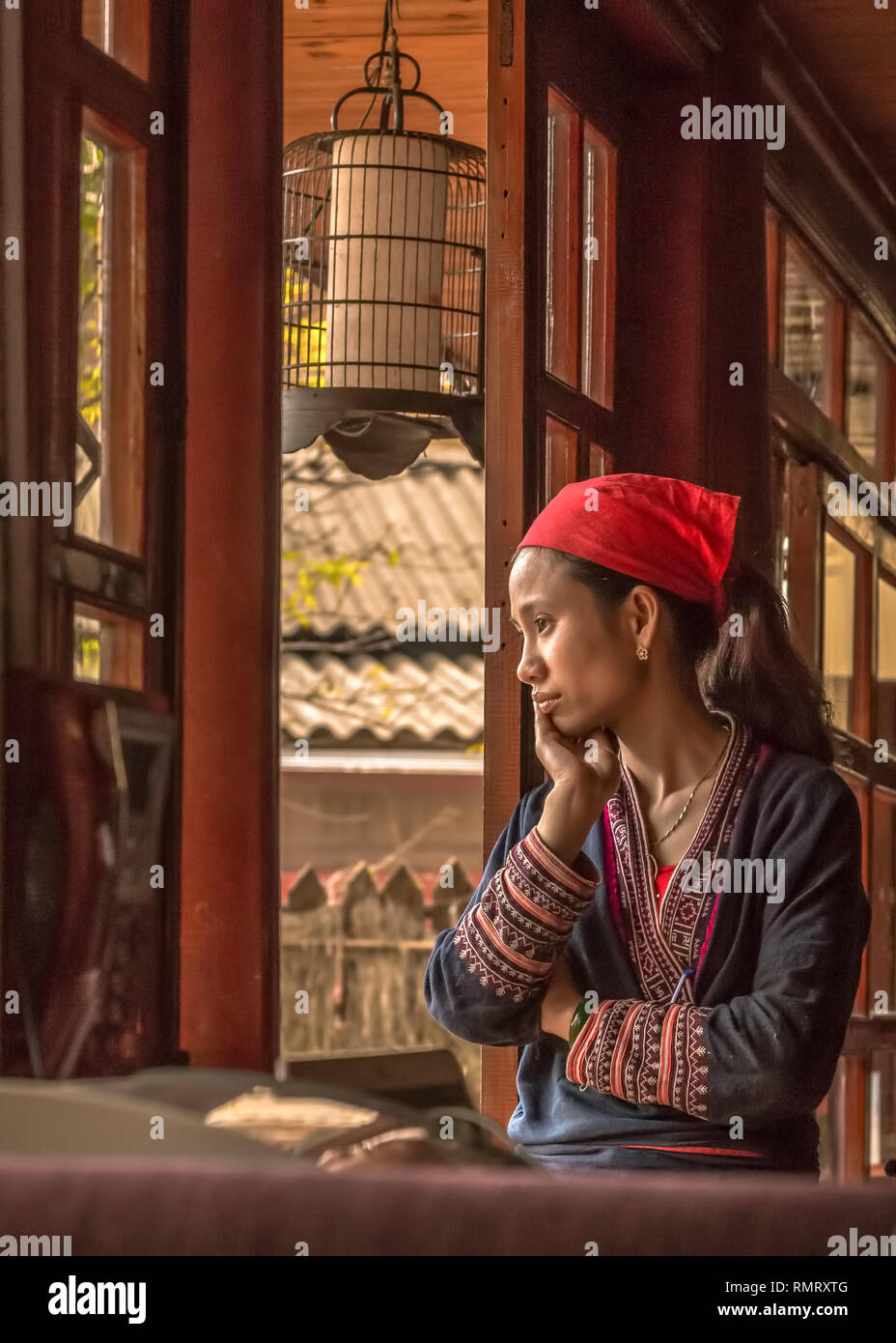 Une femme Dao rouge dans les vêtements brodés minutieusement regards par un fenêtre verticale dans un bâtiment traditionnel à Sapa, Vietnam. Banque D'Images