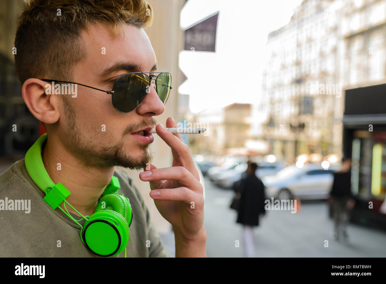 Jeune homme séduisant avec casque et lunettes vert bénéficiant d'une cigarette dans la rue. Banque D'Images