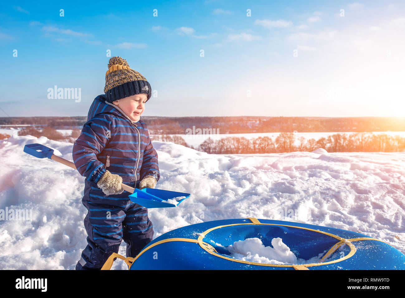 Petit garçon avec une pelle à neige en hiver dans le parc. Heureux qui peut accueillir la neige en tubes, dans des vêtements décontractés. Banque D'Images