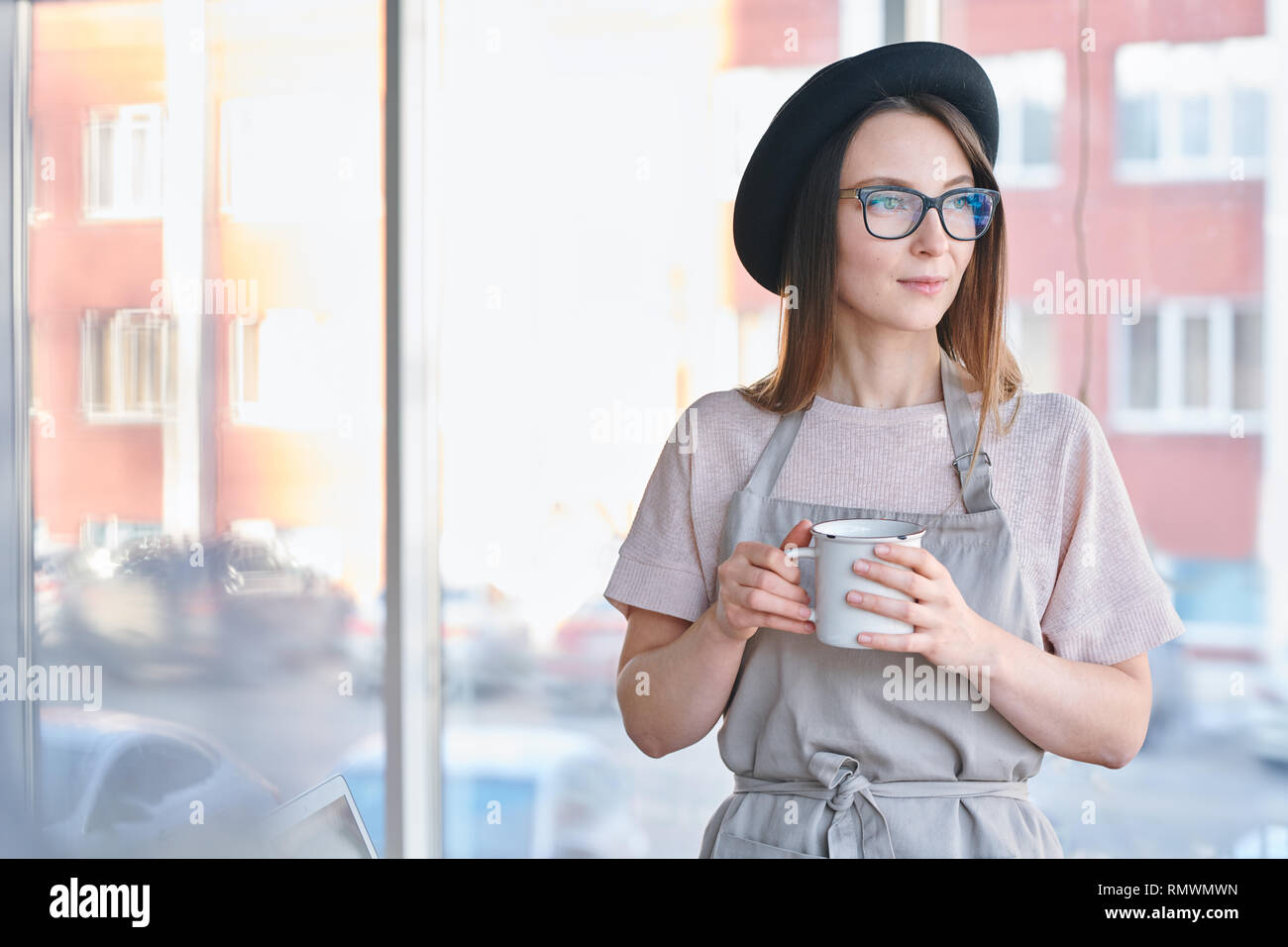 Jolie jeune femme sereine avec chapeau, lunettes et vêtements de travail prenant le thé ou café à partir de mug en studio Banque D'Images