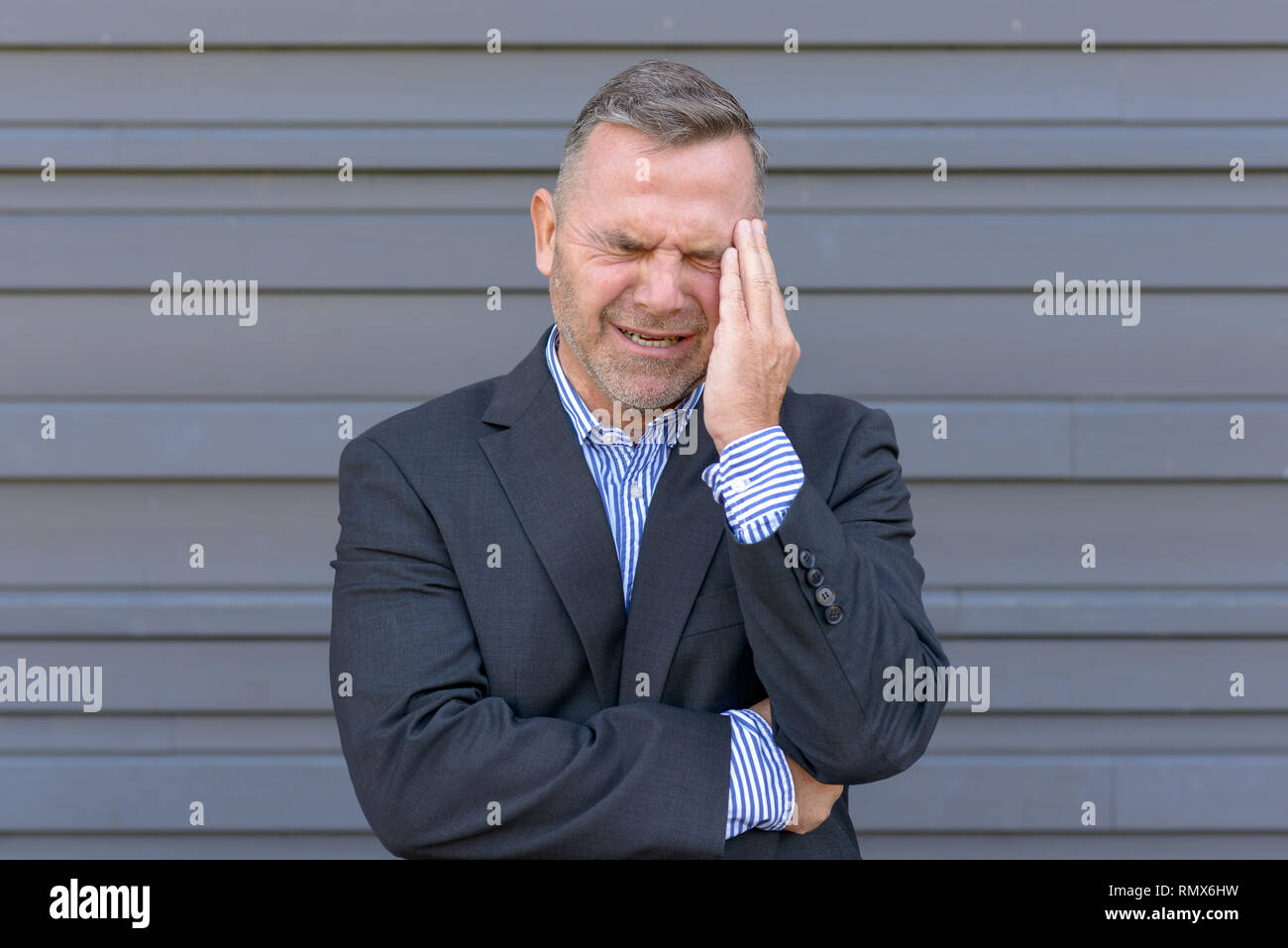 Stressed businessman, ou souffrant d'un mal de tête, debout avec sa main à son temple grimaçant contre un mur extérieur gris Banque D'Images