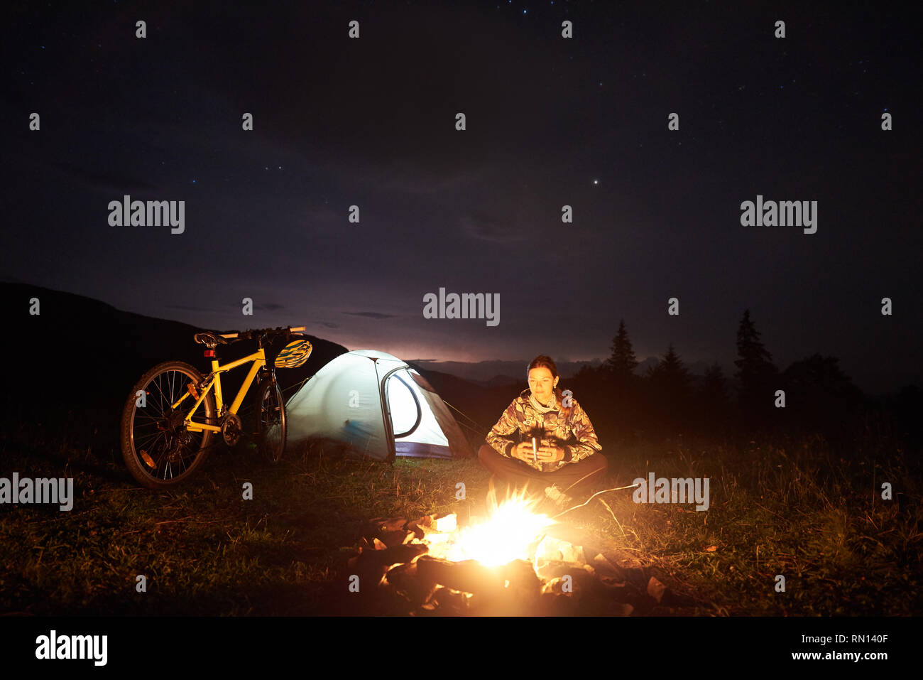 Young woman cyclist bénéficiant de nuit camping près de la gravure de camp, allumé, tente tourisme vtt sous beau soir ciel plein d'étoiles. Tourisme et activités de plein air concept Banque D'Images