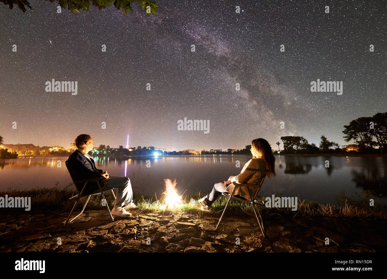 Homme et femme d'affaires reposant sur des chaises près de camp sur une rive du lac, friends enjoying soir ciel plein d'étoiles et Voie lactée au-dessus de l'eau encore et lumineux sur la ville historique. Nuit de camping Banque D'Images